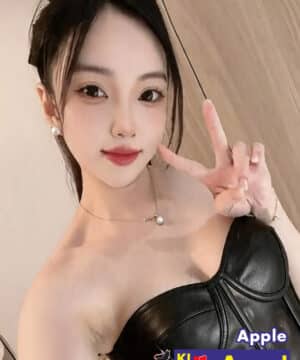 Selfie of a young woman making a peace sign, wearing a black strapless top and a pearl necklace, with an escort ad badge in the corner.